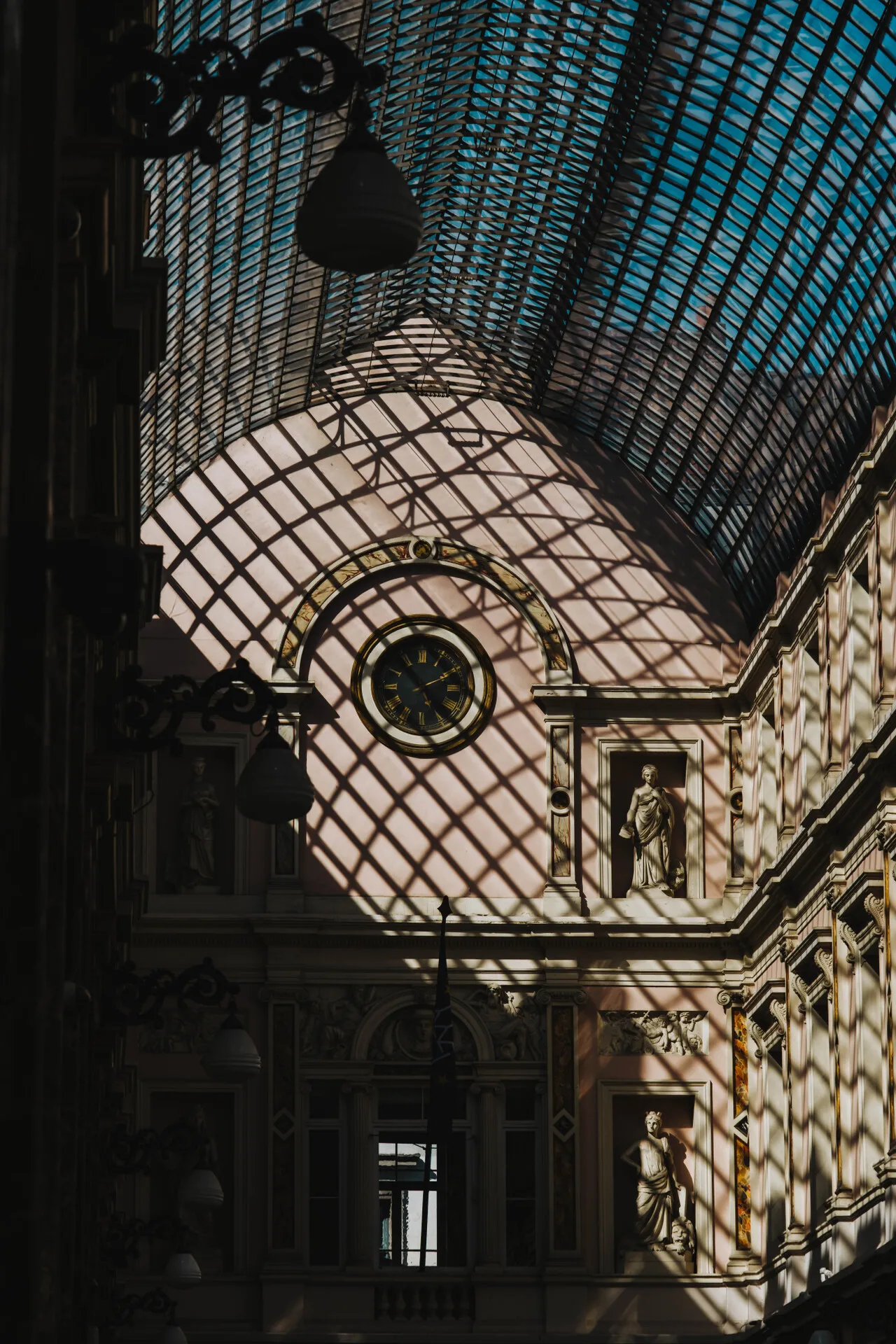 Large arcades in a building with a glass roof. The roof also has a quadratic pattern of metal rods, and the sun is projecting shadows of that rods to the inside of the building.